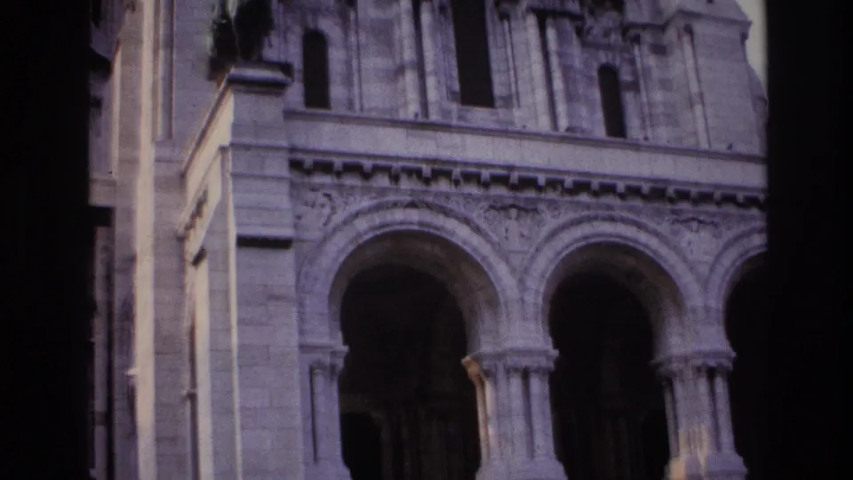 Handheld 1967 Paris frame centered on two arched portals, heavy columns, carved stonework, and shadowed entrance doors.