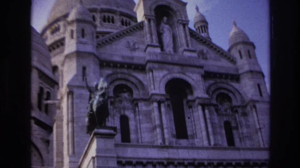 1967 Paris home-movie frame showing Sacre-Coeur turrets, facade sculpture, arched openings, and a bronze statue.