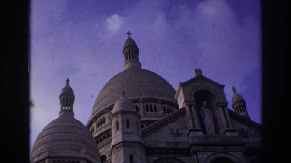 1967 8mm Paris still with Sacre-Coeur's rounded domes, entrance sculpture, and ornate stonework under a lightly clouded sky.