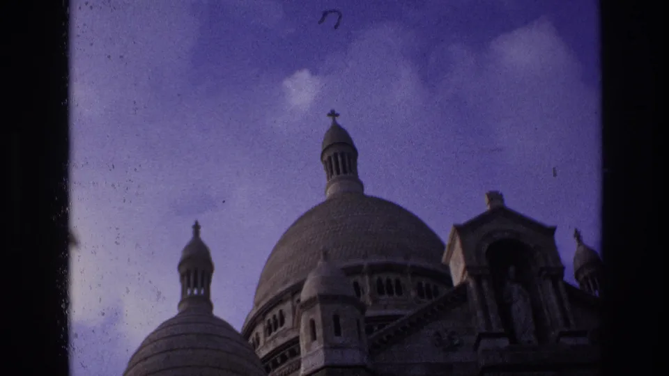 1967 Paris home-movie frame showing Sacre-Coeur's domes and facade in darker exposure, bordered by the 8mm frame edge.