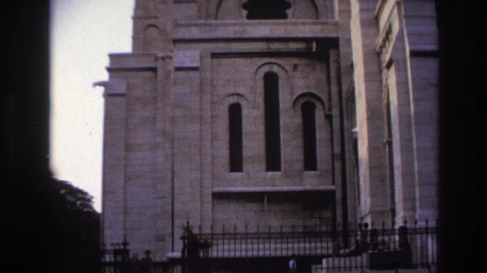 1967 Paris frame showing Sacre-Coeur's lower stone facade, tall arched windows, iron fencing, and small distant visitors at ground level.