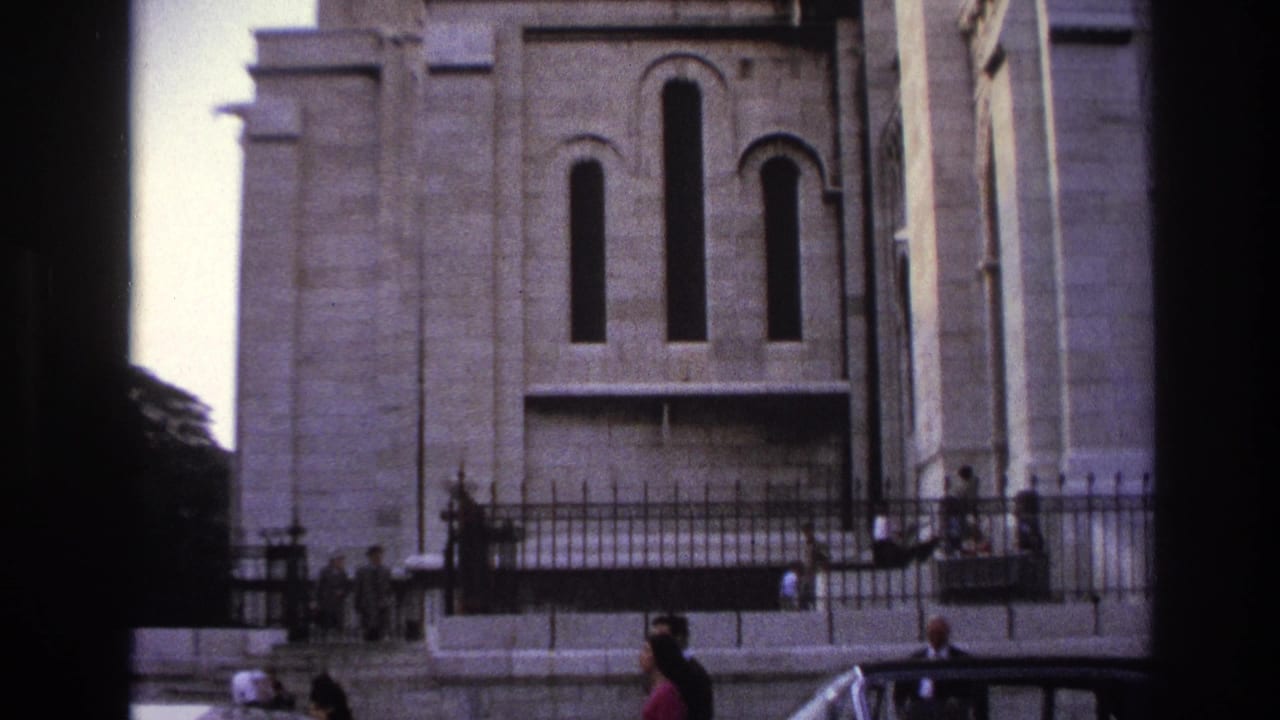 1967 Paris home-movie frame showing a grand stone facade, wrought-iron fence, pedestrians on the sidewalk, and a dark sedan passing below.