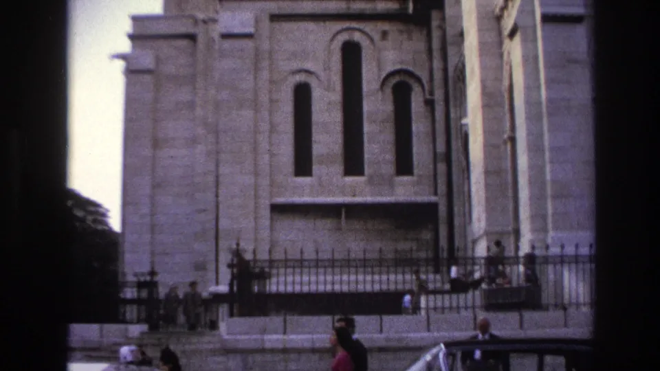 1967 Paris home-movie frame showing a grand stone facade, wrought-iron fence, pedestrians on the sidewalk, and a dark sedan passing below.