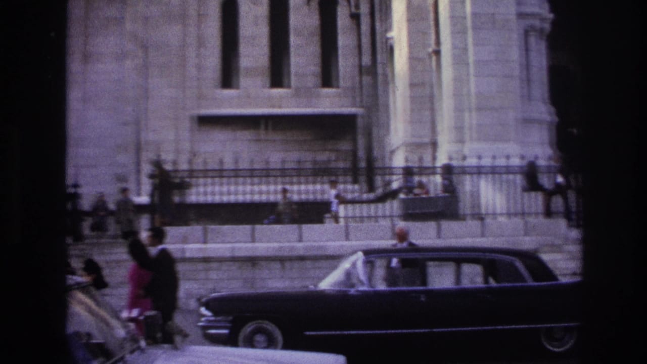 Dark sedan passing a stone building while pedestrians stand near an iron fence.