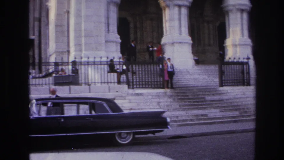 1967 Paris frame showing broad stone steps, pedestrians near a gated entrance, and a dark sedan moving along the street below.