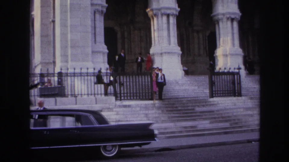 1967 Paris home-movie view of pedestrians on broad steps beside a wrought-iron gate, with a dark sedan passing in the foreground.