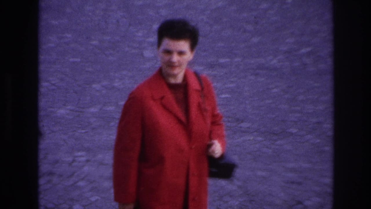 In 1967 Paris home-movie footage, a woman in a bright red coat walks toward the camera across cobblestones while carrying a black handbag.