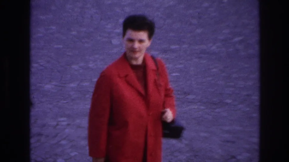 In 1967 Paris home-movie footage, a woman in a bright red coat walks toward the camera across cobblestones while carrying a black handbag.