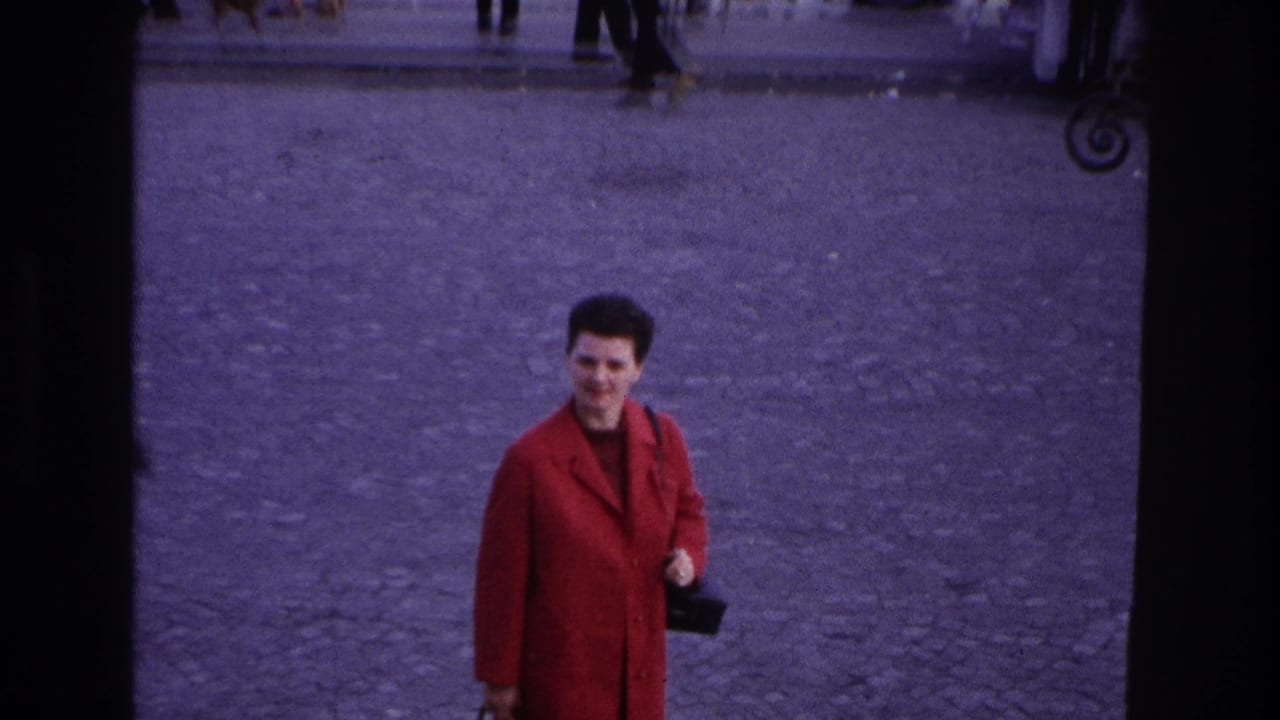 The woman in the red coat appears farther from the camera on a Paris cobblestone plaza, smiling or looking toward the person filming.
