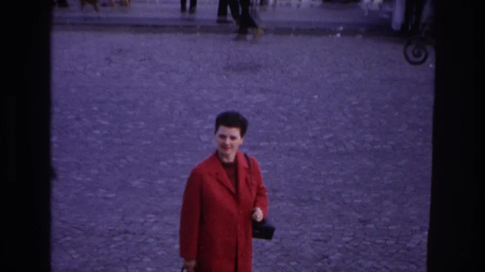 The woman in the red coat appears farther from the camera on a Paris cobblestone plaza, smiling or looking toward the person filming.