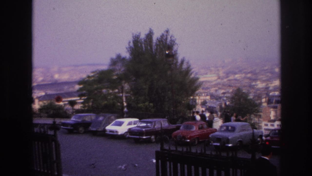 1967 Paris home-movie frame showing parked cars along an overlook road, dark railings, trees, pedestrians, and a hazy city panorama.