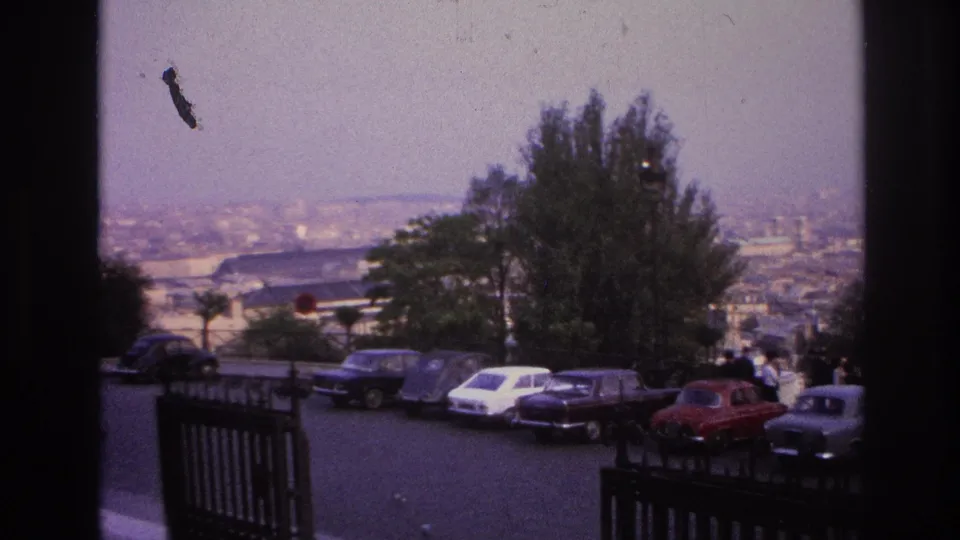 1967 Paris overlook view with compact cars parked along the road, people near the railing, trees, and a hazy city skyline in the distance.