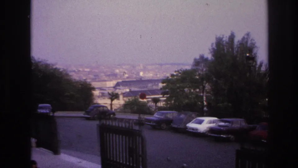 1967 Paris home-movie frame looking across the overlook road toward parked cars, dark railings, trees, and a soft panoramic city view.