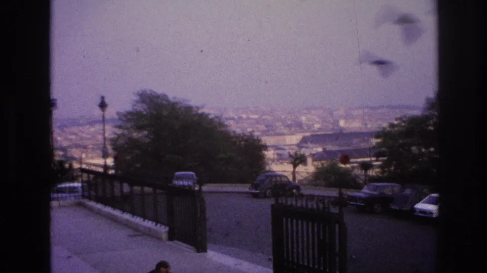 1967 Paris frame from a scenic overlook, with a railing, parked cars, trees, a few distant people, and the city receding under a muted sky.