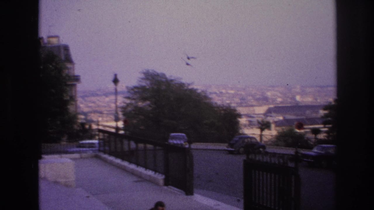 1967 Paris home-movie frame looking through a dark edge toward a sloped overlook road, iron fence, trees, cars, and distant rooftops.