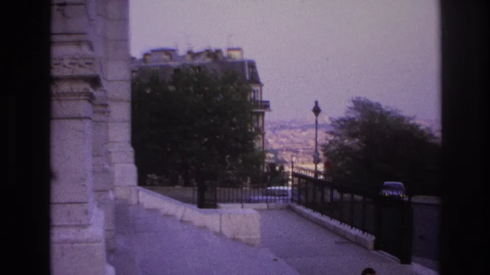 1967 Paris frame with a stone structure at left, steps and railing below, trees, cars, and hazy rooftops beyond the overlook.