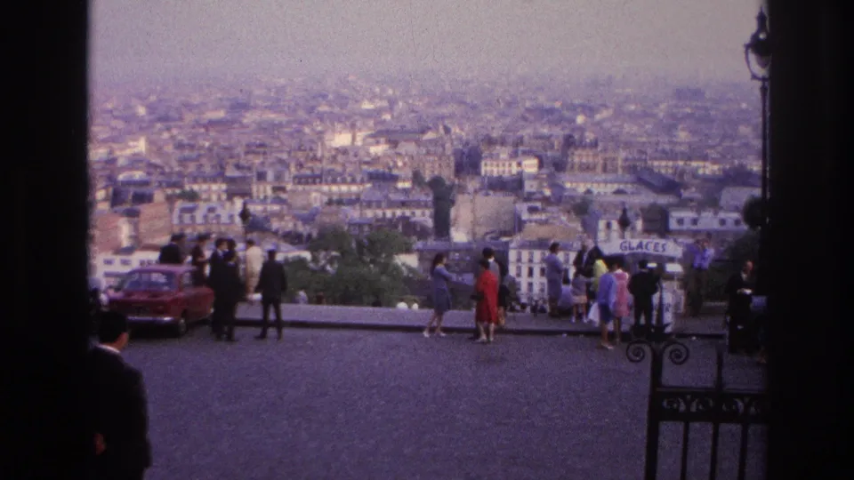 1967 Paris overlook scene with visitors near the railing, a red car, dense rooftops, hazy sky, and a small GLACES sign at right.