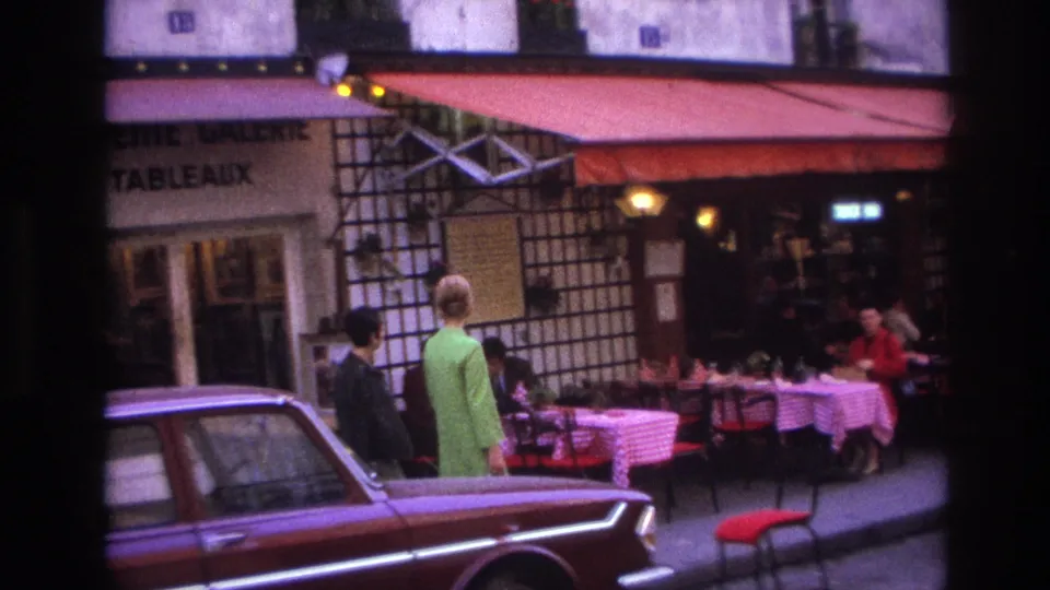 1967 Paris restaurant frontage with two pedestrians beside outdoor seating, diners at checkered tables, red awnings, and a maroon car along the curb.