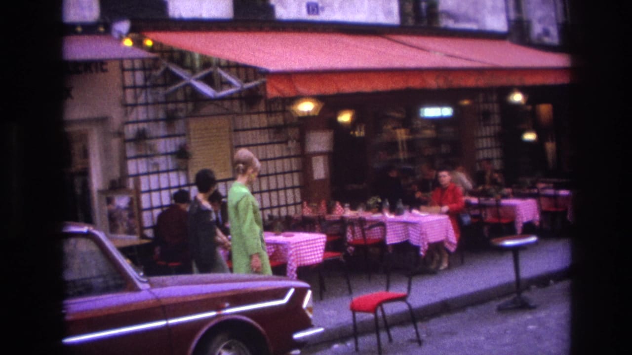 In 1967 Paris, two women pass a sidewalk cafe with red-and-white tablecloths, a red awning, diners, and parked cars.