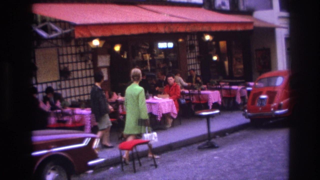 Women approach cafe tables as diners sit under a red awning beside a red car.
