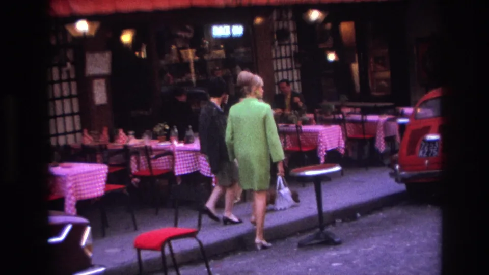 Two women, one in a green coat, walk away along a Paris street beside outdoor cafe seating and mid-century parked cars.