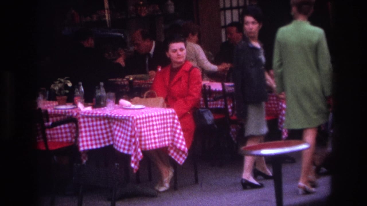 In 1967 Paris home-movie footage, a woman in a red coat sits beside red-and-white checkered cafe tables as people move through the scene.