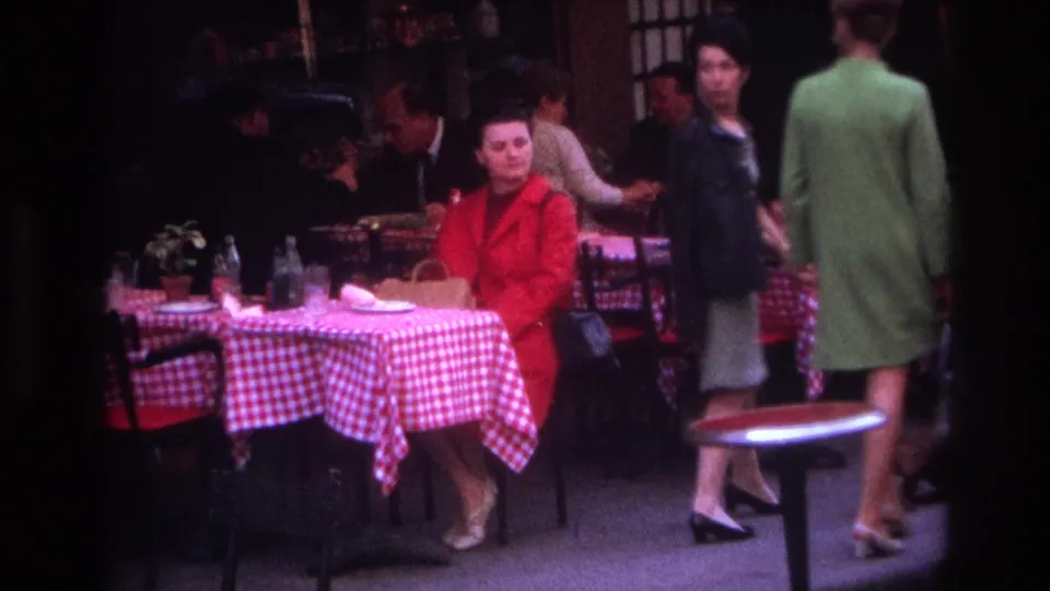 In 1967 Paris home-movie footage, a woman in a red coat sits beside red-and-white checkered cafe tables as people move through the scene.