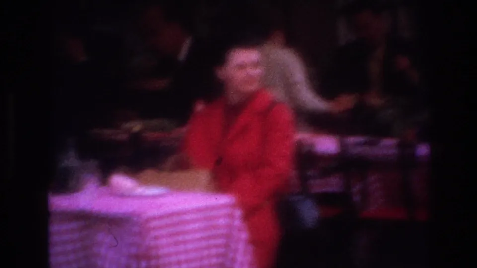 The home-movie camera catches a blurred close view of the woman in red beside a checkered table, with background figures in low light.