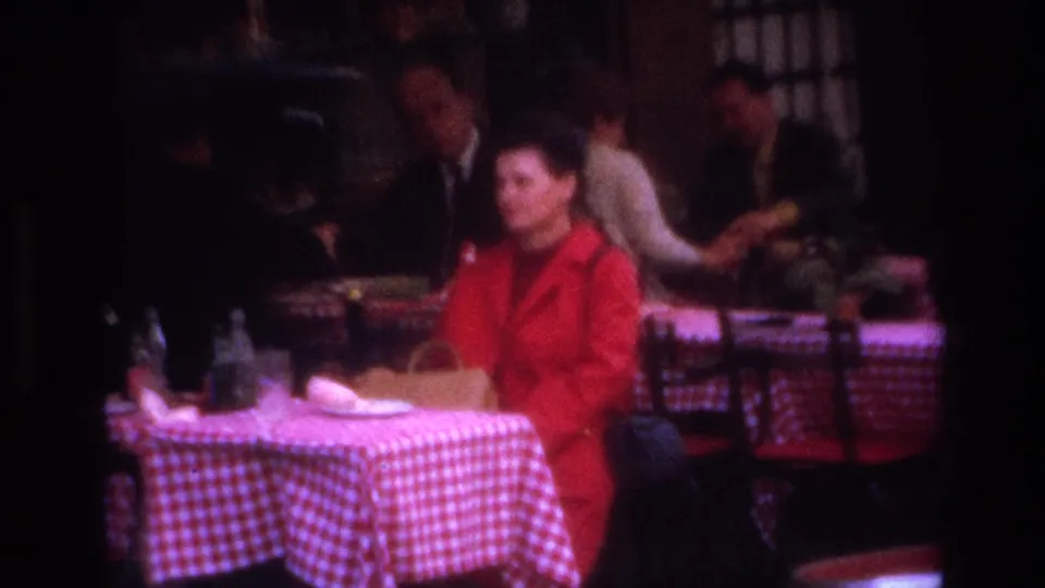 Paris cafe frame with the red-coated woman seated near bottles and a plate on a checkered table, while bystanders occupy the dim background.