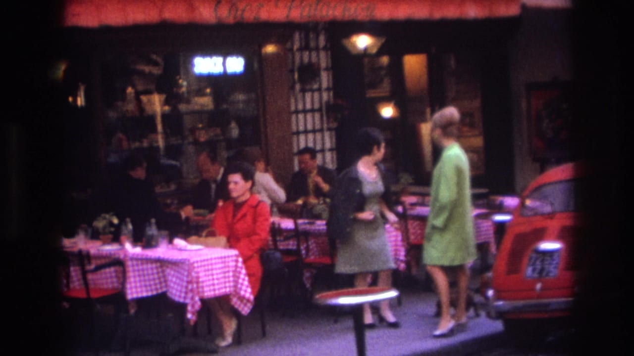 1967 Paris sidewalk cafe with pedestrians in period clothing passing seated diners beneath a storefront awning.