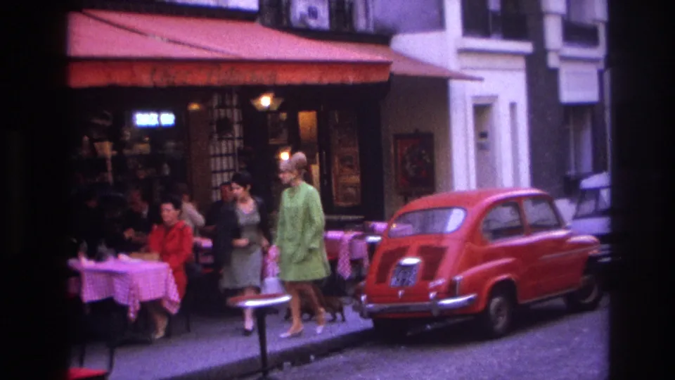 1967 Paris street view showing an outdoor cafe, passing pedestrians, and a red compact car parked beside the curb.