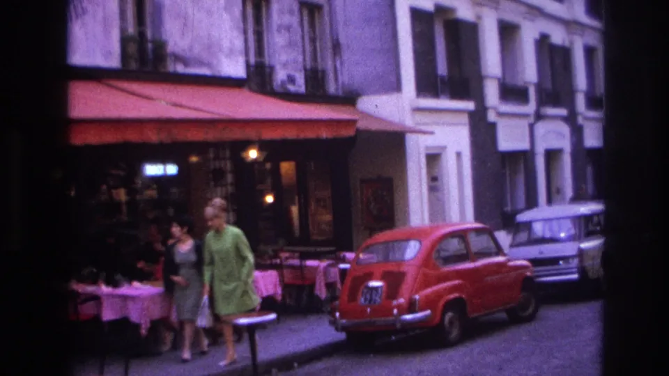 Wider 1967 Paris street frame with a sidewalk cafe, pedestrians, a red compact car, and another parked car beyond it.