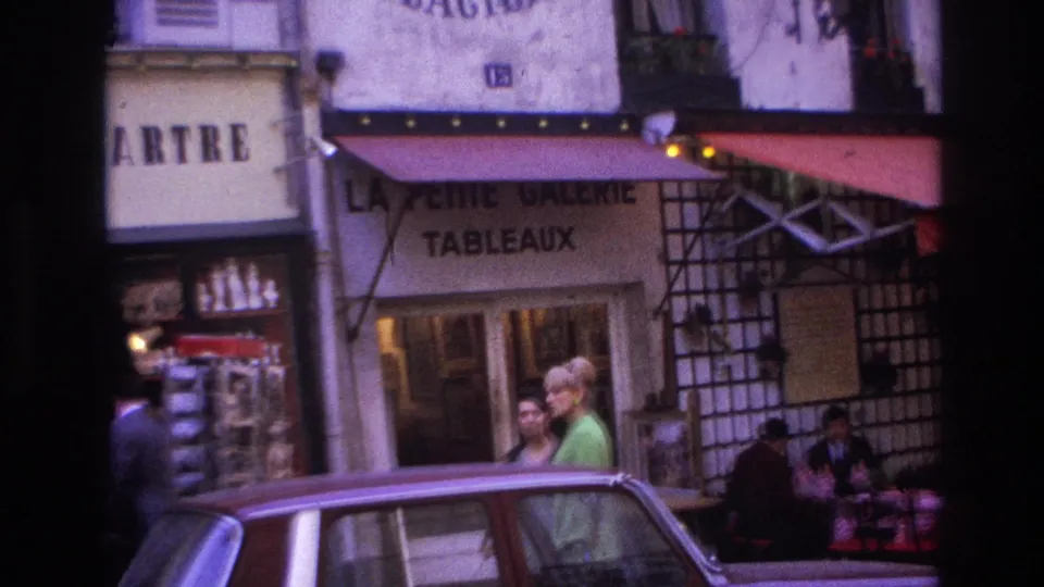1967 Paris street view with cafe awnings, storefront lettering, pedestrians near the sidewalk, and a car in the foreground.