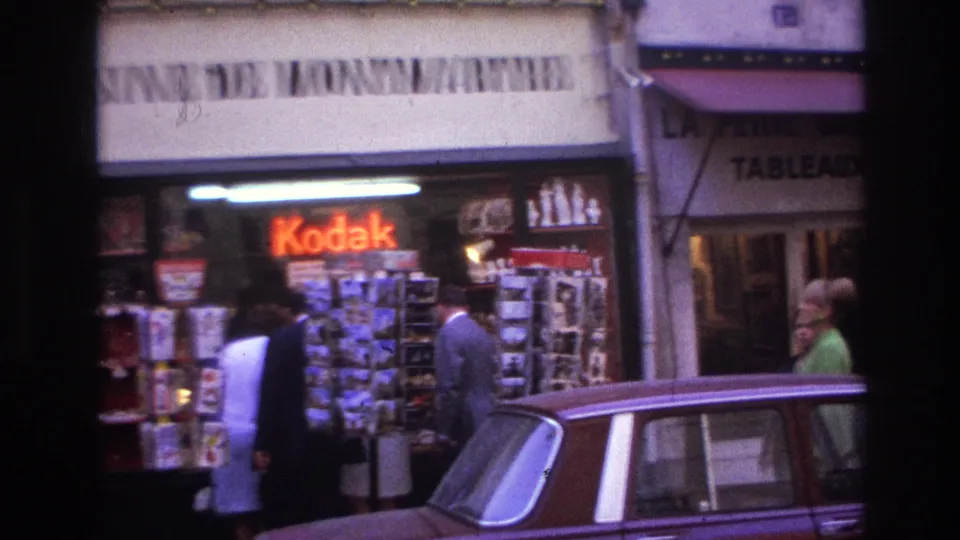 1967 Paris frame showing a Kodak sign, outdoor postcard racks, pedestrians at the shopfront, and a car in front.