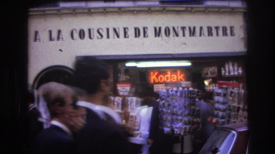 1967 Paris street frame with large French facade lettering, a Kodak storefront, postcard racks, pedestrians, and a car edge.