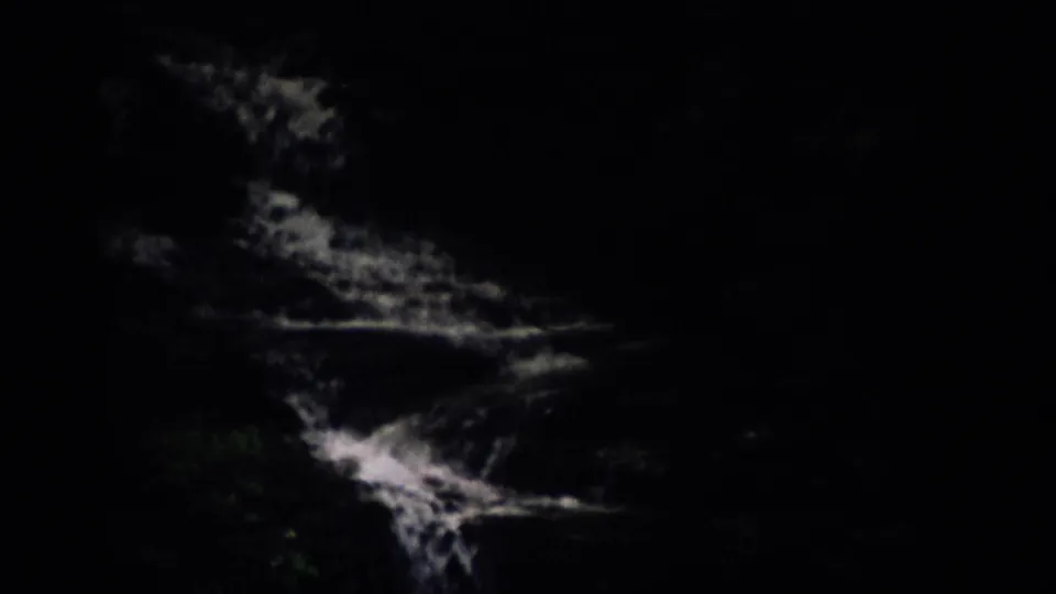 Underexposed 8mm frame of water moving through a shaded wooded stream or falls area, with trees faintly visible around it.