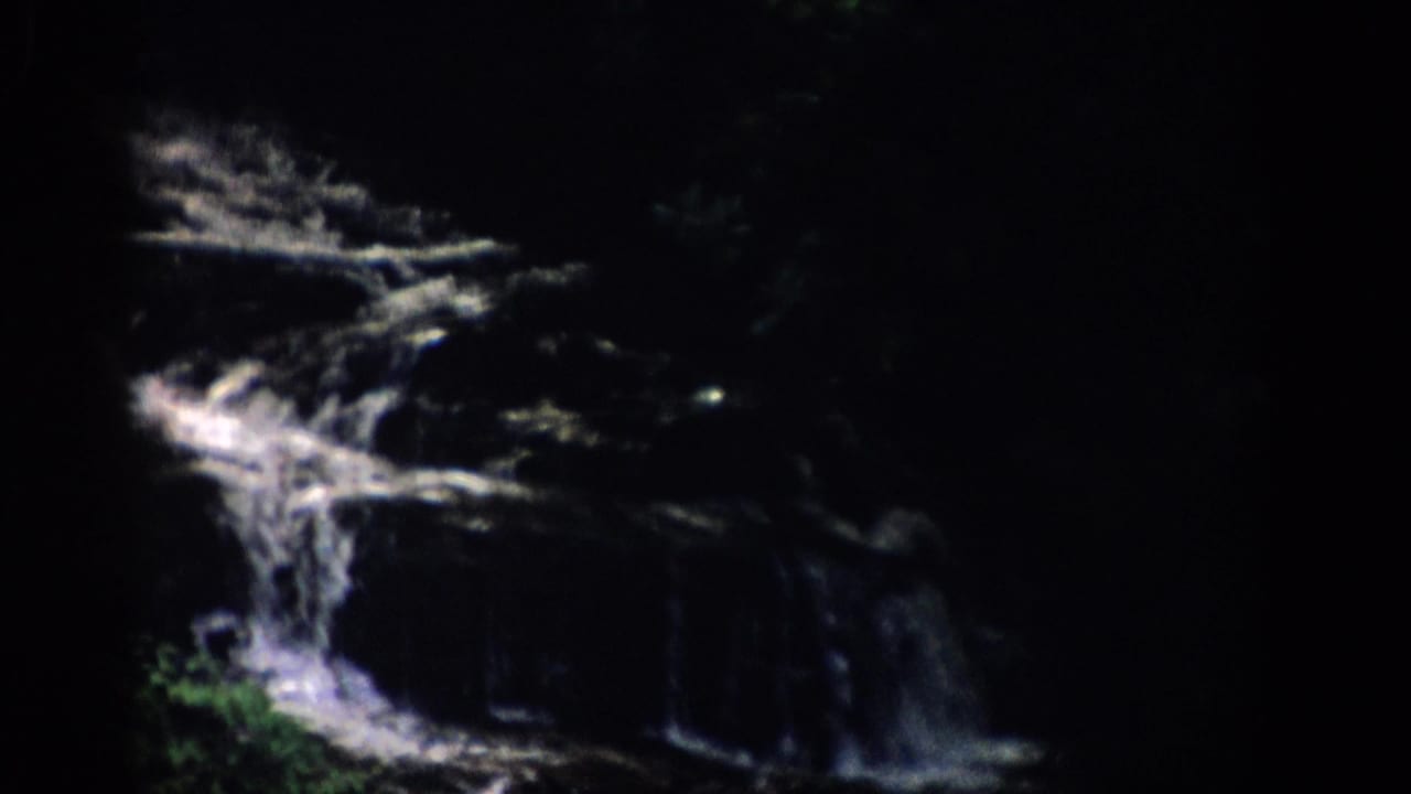 Waterfall descending across dark rock ledges with foliage near the lower edge.