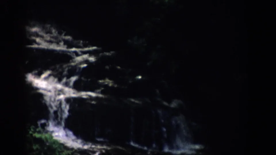 1959 Colorado home-movie frame of a small waterfall crossing shadowed rock, with green leaves visible near the base.
