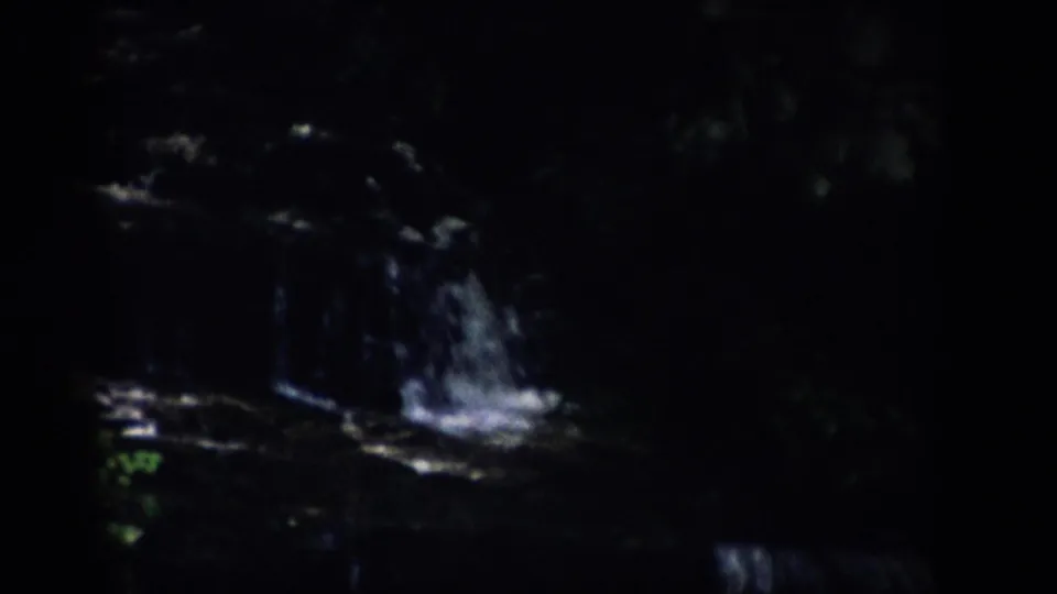 1959 Colorado home-movie frame with a dimly lit waterfall dropping over dark rock shelves in a forested recess.