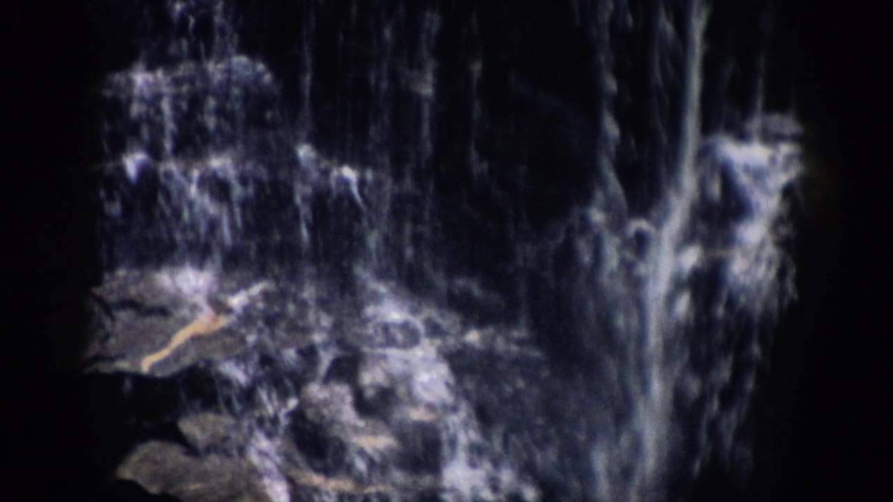 Colorado 1959 home-movie frame showing a shadowed waterfall flowing over rocks, with soft 8mm grain and dark frame edges.