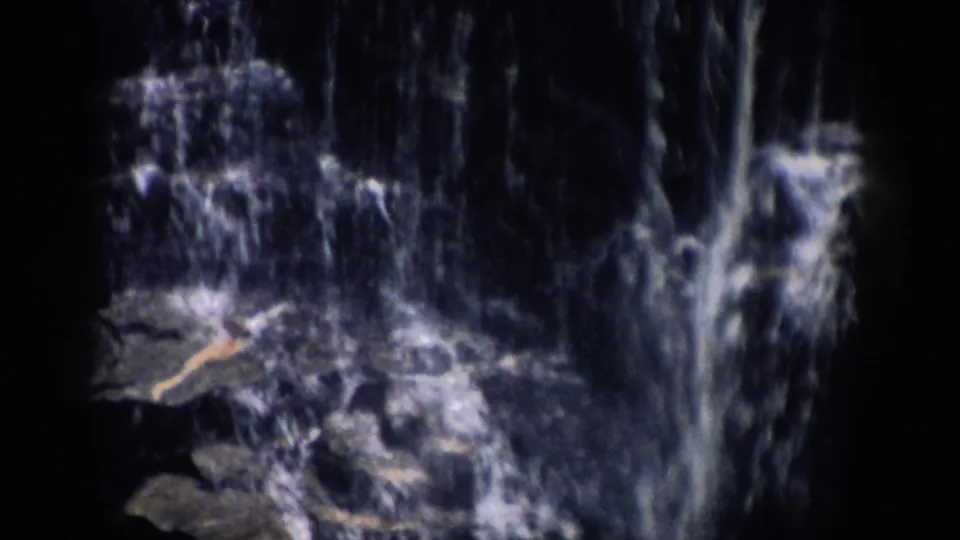 Colorado 1959 home-movie frame showing a shadowed waterfall flowing over rocks, with soft 8mm grain and dark frame edges.