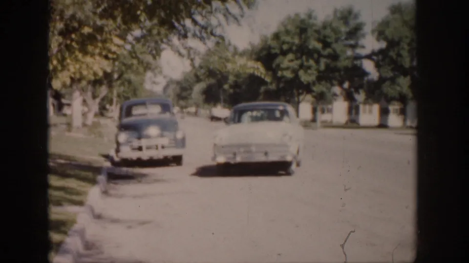 Colorado 1959 home-movie frame with two late-1950s cars on a bright, tree-lined street with lawns and houses in the distance.