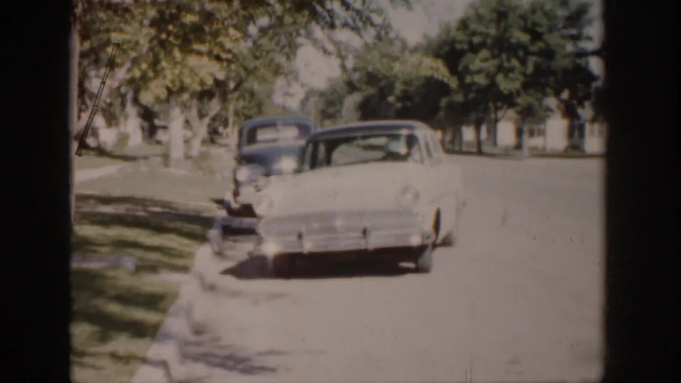 Colorado 1959 home-movie frame showing a white mid-century sedan approaching on a leafy street, with another dark car behind it.
