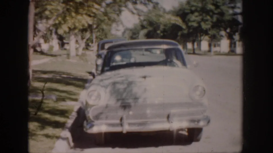 Colorado 1959 home-movie frame with a white sedan close to camera, dark car behind, trees, curb, and bright residential street light.