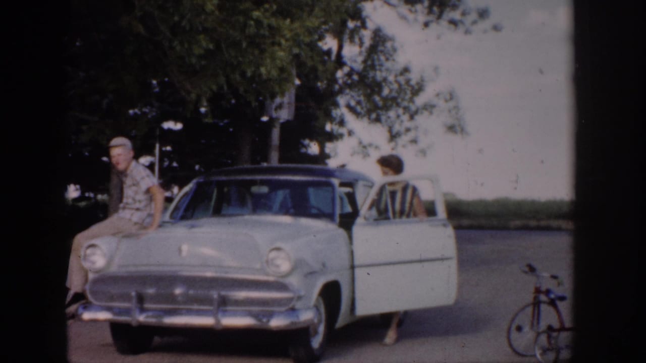 1959 Colorado 8mm frame showing a man on a light sedan hood, a woman near the open door, a bicycle, trees, and a house edge.