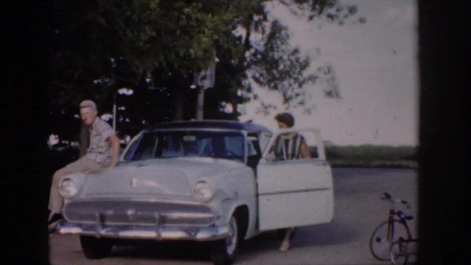 1959 Colorado 8mm frame showing a man on a light sedan hood, a woman near the open door, a bicycle, trees, and a house edge.