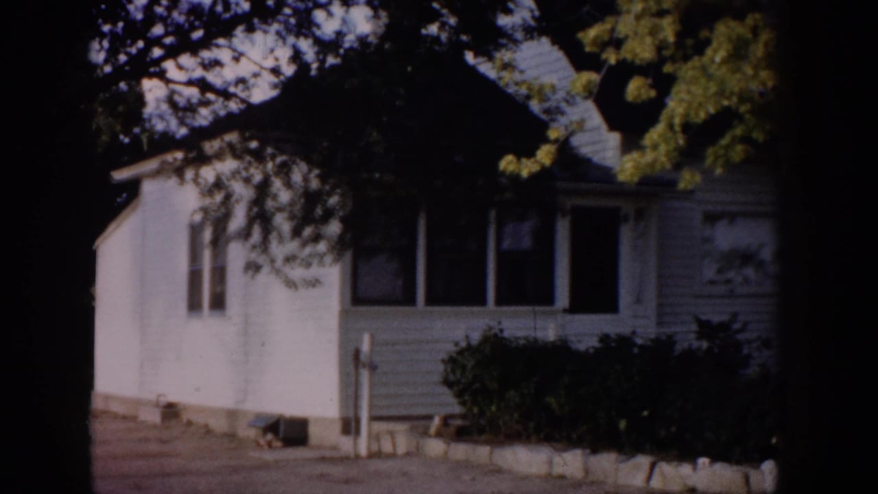 White clapboard house exterior under tree shade, with driveway and shrubs visible.