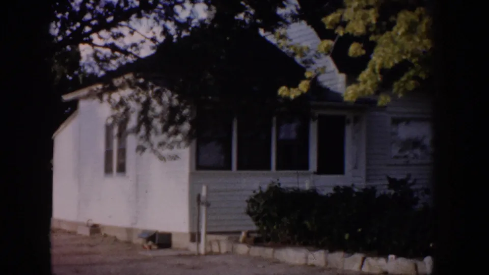1959 Colorado home-movie frame showing a white clapboard house exterior, shaded windows, shrubs, driveway, and tree branches.