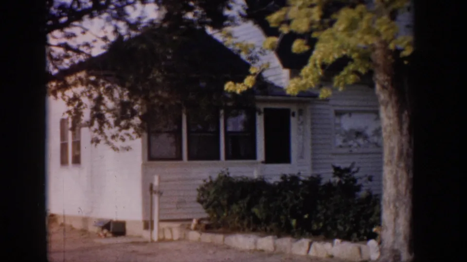 1959 Colorado 8mm frame of a small house exterior with clapboard siding, shrubs, tree trunk, stone edging, and driveway.