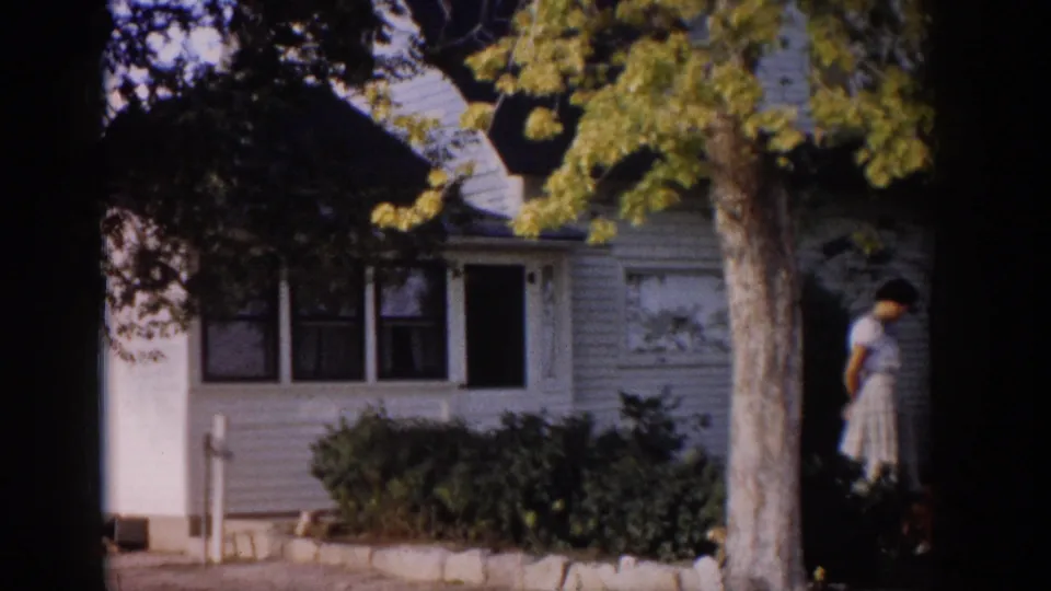 1959 Colorado home-movie frame showing a woman in a light dress near the side of a clapboard house, with shrubs and a tree in front.